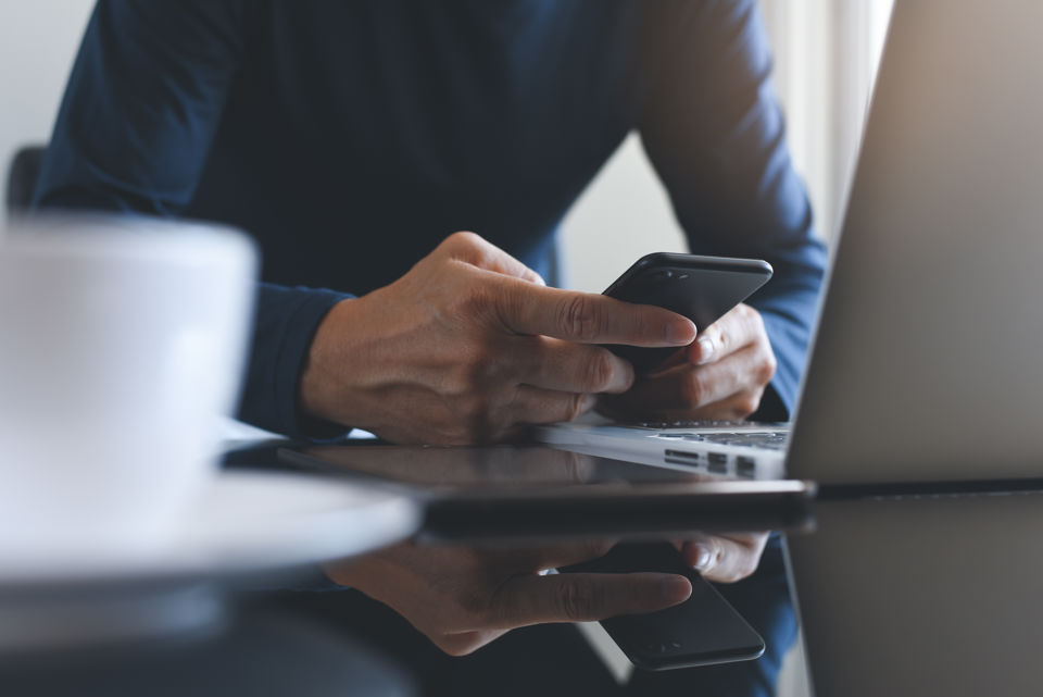 Eine Person sitzt vor dem Notebook und tippt etwas auf dem Smartphone ein