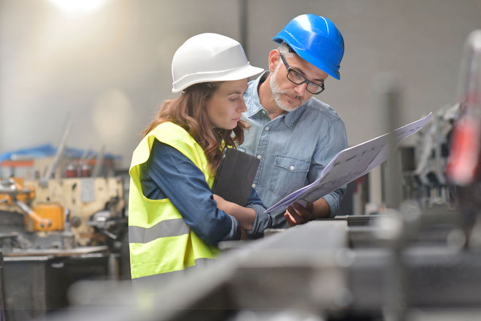 Symboldbild für Wirtschaft der Industrie: Eine Frau und ein Mann mit Helmen in einer Werkhalle 