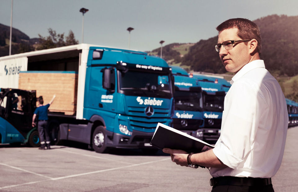 Ein Mann in weissem Hemd steht auf einem Parkplatz mit vielen LKW und beobachtet die Beladung der LKW