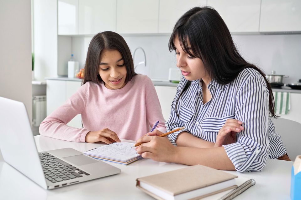 Eine Mutter sitzt mit ihrer Tochter am Esstisch und sie erstellen zusammen in einem Notizbuch eine Budgetaufstellung